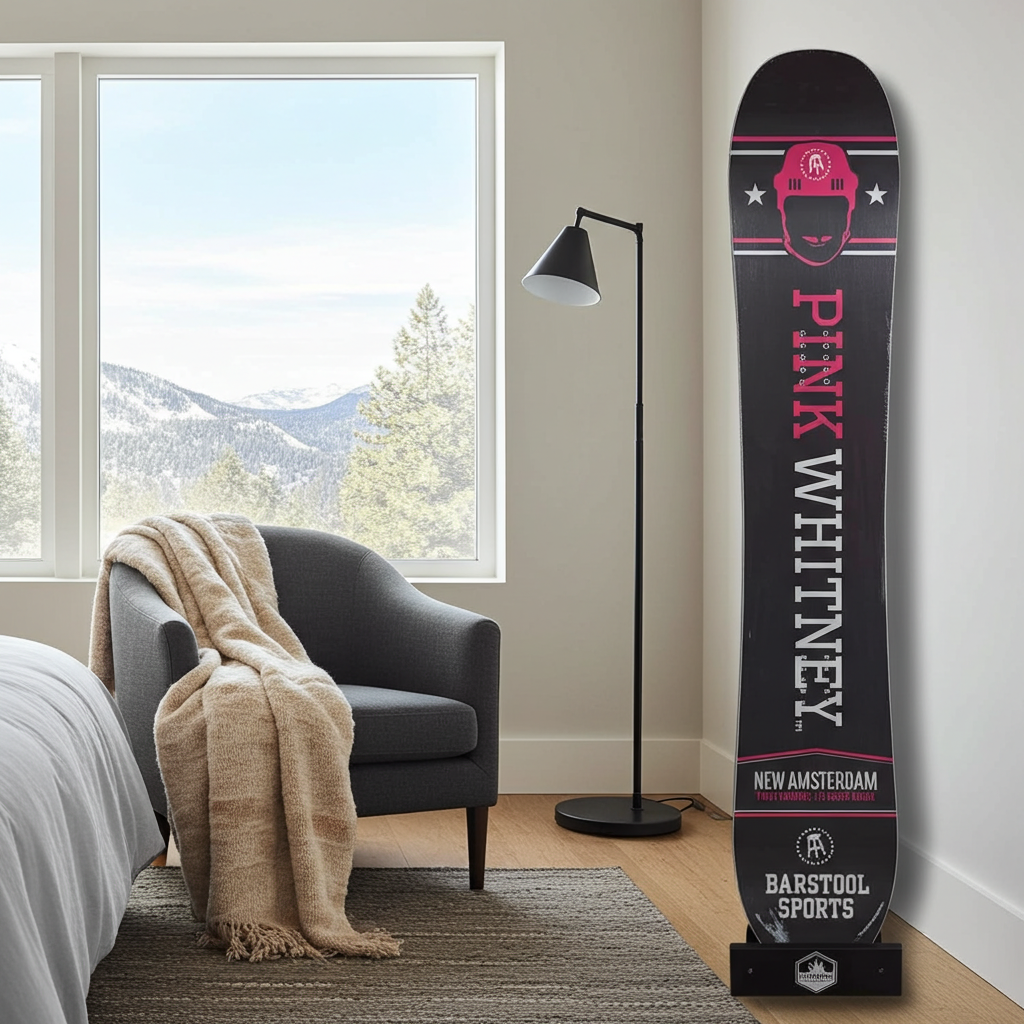 A single snowboard standing upright in a bedroom corner beside a window, chair, floor lamp, and bed. The board is predominantly black with bold pink text and graphics, plus small white star accents.